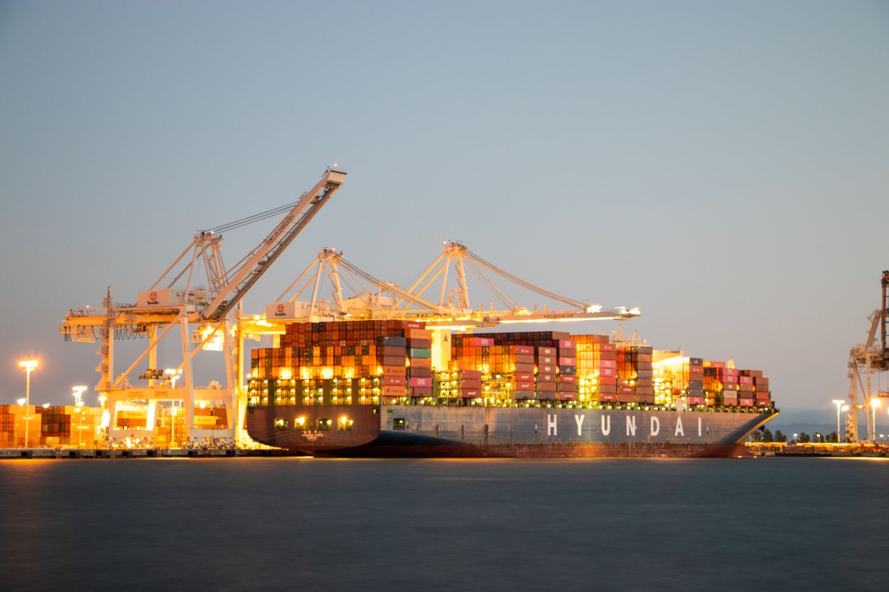 why-choose-us Container ship docked at port under cranes during twilight, illuminated against the evening sky.